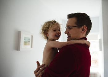 Father carrying happy daughter while standing by wall at home