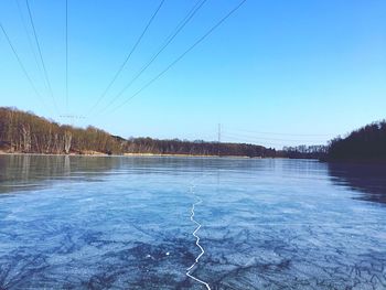 Power lines hanging over lake against clear blue sky