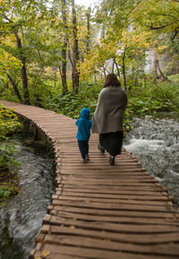 Rear view of mother and son walking on boardwalk over stream in forest