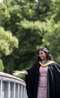 Smiling young woman wearing graduation gown standing against trees