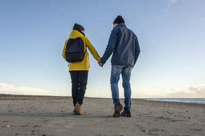 Rear view of a mature couple walking at beach holding hands, backpack and yellow coat, fall.