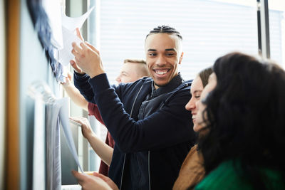 Happy young man with female friends checking papers on bulletin board at university