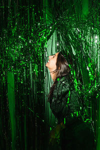 Delighted female wearing festive dress playing with green shiny tinsel stripes at party while looking at camera