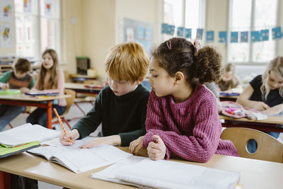 Girl sitting besides blond boy writing in book at desk in classroom