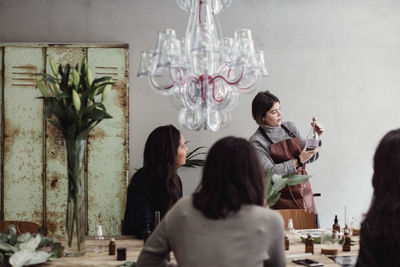 Mid adult female owner showing perfume bottle to colleagues sitting at workshop