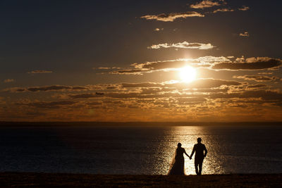 Silhouette couple holding hands while standing at beach against sky during sunset