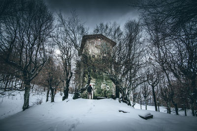 Bare trees and rurale building on snow covered field against sky