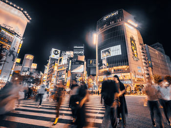 Blurred motion of people walking on road against illuminated buildings at night