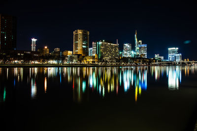 Illuminated buildings by river against sky at night