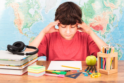 Stressed boy sitting with books on desk against map