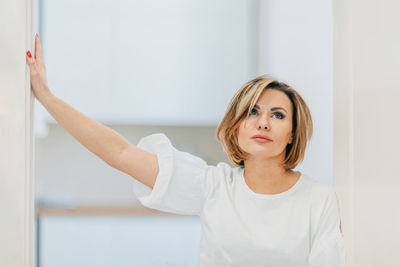 An adult, beautiful, well-groomed woman stands with a dreamy look and a slight smile in the doorway