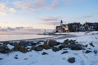 Snow covered land by sea against sky during sunset