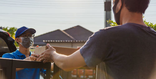 Rear view of man wearing mask taking parcel from delivery person outdoors