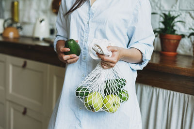 Young woman in shirt hold knitted rag bag shop with avocado and green apples in hands on kitchen