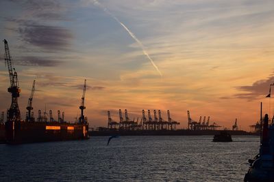 Cranes at commercial dock against sky during sunset