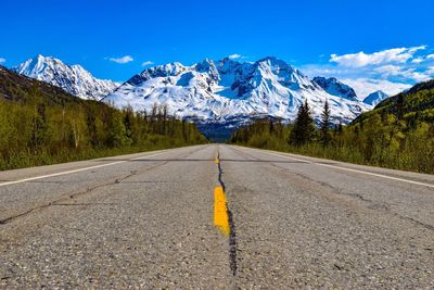 Empty road leading towards snowcapped mountains against blue sky