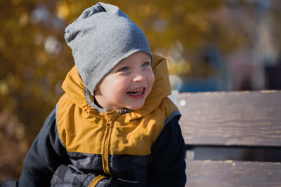 Happy and laughing child on a wooden bench. a small boy in a jacket and hat rests in a park