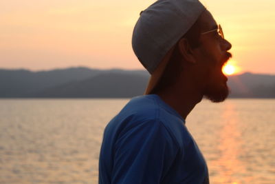 Side view of young man standing by sea against sky during sunset