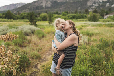 Happy mother kissing cute daughter while standing on grassy landscape against mountains in forest