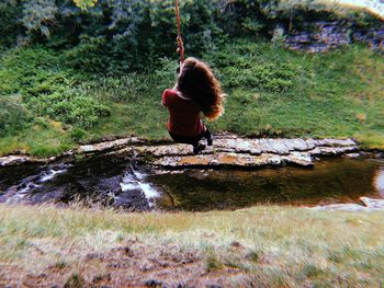 Rear view of woman hanging from rope swing by stream