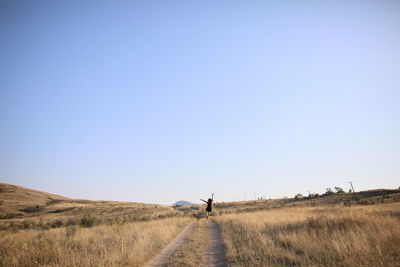 Rear view of woman on dirt road amidst grassy landscape against clear sky