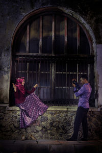 Young man photographing smiling girlfriend sitting on building wall at dusk