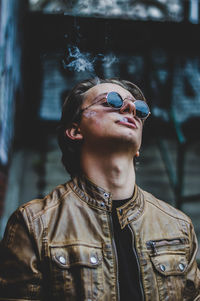 Young man wearing sunglasses and leather jacket while emitting smoke from mouth