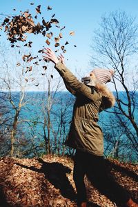 Side view of woman throwing dried leaves on hill by sea against sky