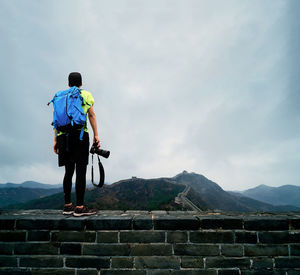 Rear view of backpacker standing on retaining wall against cloudy sky