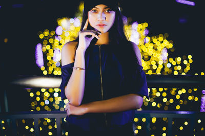Portrait of beautiful woman standing by railing against illuminated lights