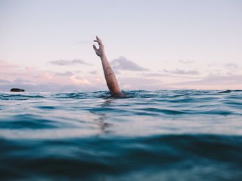 Cropped image of woman hand drowning in sea against sky