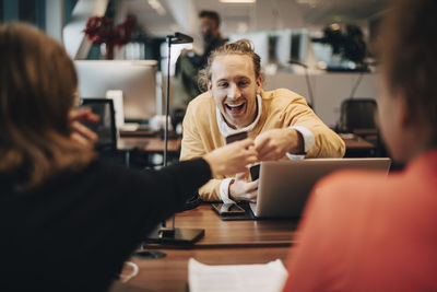 Rear view of businesswoman sitting by colleague showing smart phone to cheerful businessman in office