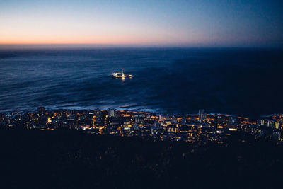 Aerial view of illuminated buildings by sea against sky at sunset