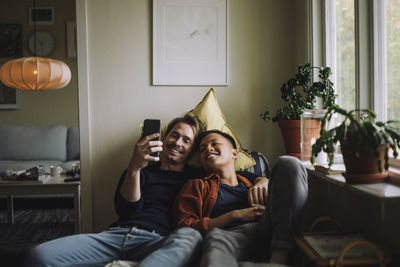 Smiling gay couple sharing smart phone while sitting near window at home