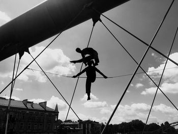 Low angle view of silhouette statues hanging from bridge cables against sky