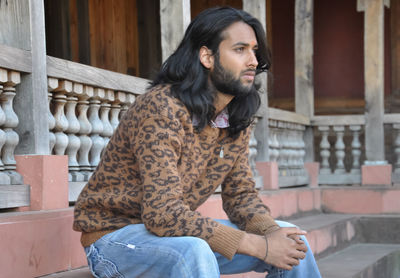 A good looking long haired indian young guy, looking sideways while sitting in temple stairs