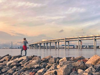 Rear view of man standing against bridge over sea during sunset