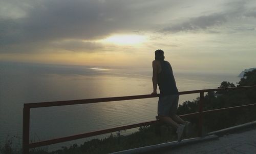 Rear view of man leaning on railing over sea against sky during sunset