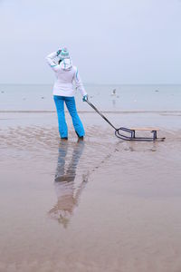 Rear view of woman holding sled while standing on shore at beach against sky