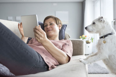 Portrait of smiling woman lying on the couch using tablet while dog watching her