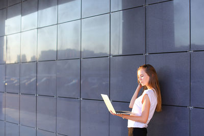 Young businesswoman using laptop standing by wall