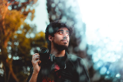 Low angle view of young man holding camera while standing in forest