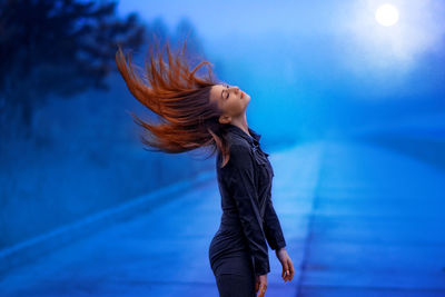 Side view of woman tossing hair while standing on road against sky at dusk