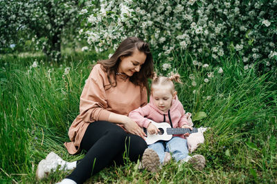 Young mother and baby daughter play the ukulele in a blooming apple orchard in nature