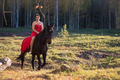 A young woman wearing a red dress rides a horse in the evening in the soft sunlight