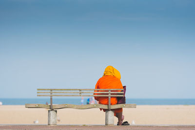 Rear view of monk sitting on bench at beach against sky