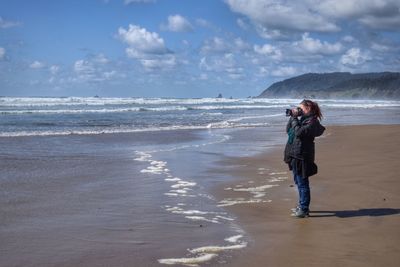 Full length side view of woman photographing at beach against sky on sunny day