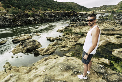 Side view of mid adult man wearing sunglasses standing on rock at riverbank