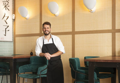 Cheerful male chef in apron standing in sushi restaurant and looking at camera