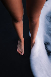 From above of crop unrecognizable barefoot female traveler standing on wet sandy shore washed by foamy wave